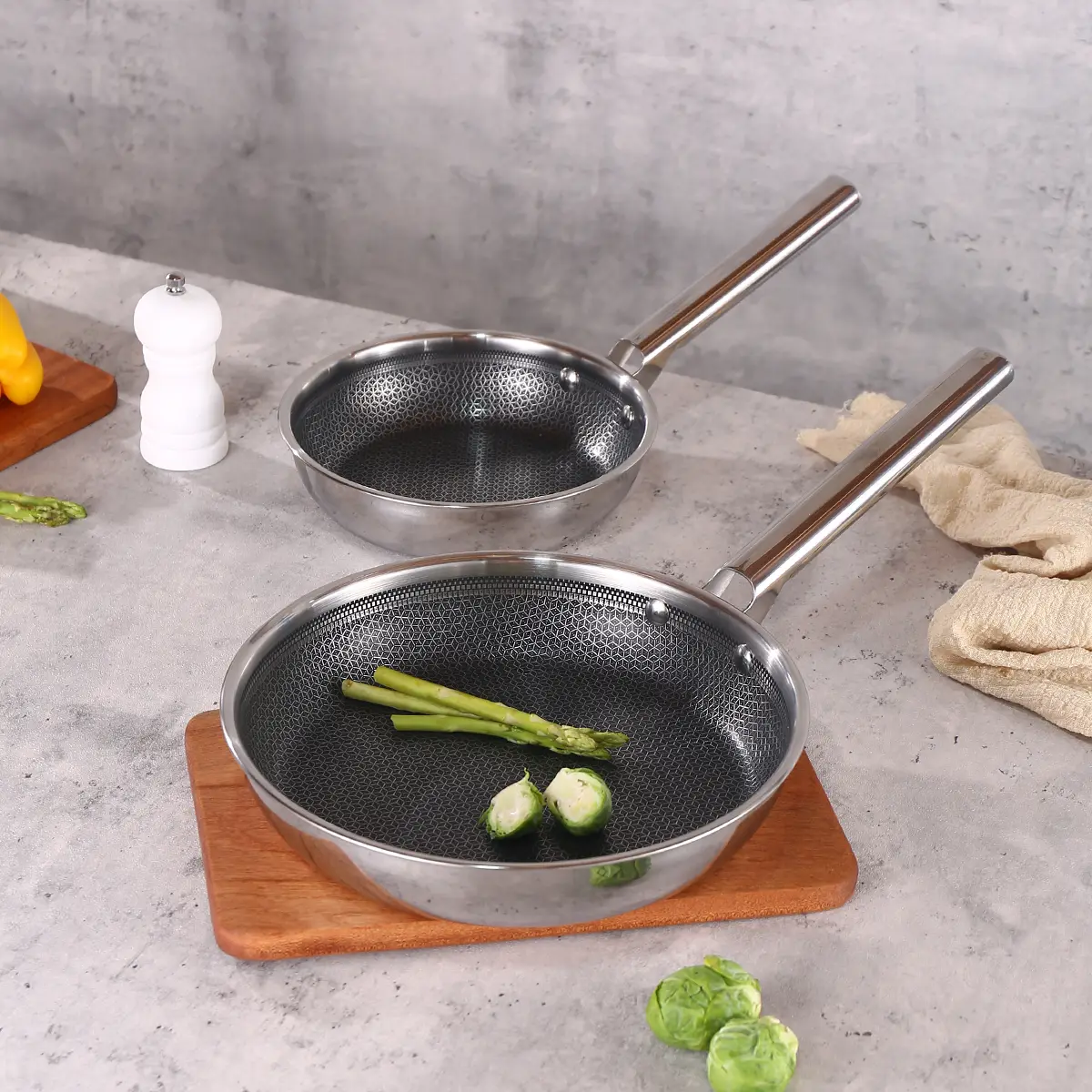 A wooden cutting board holds two stainless steel frying pans, highlighting their polished finish and modern appearance.