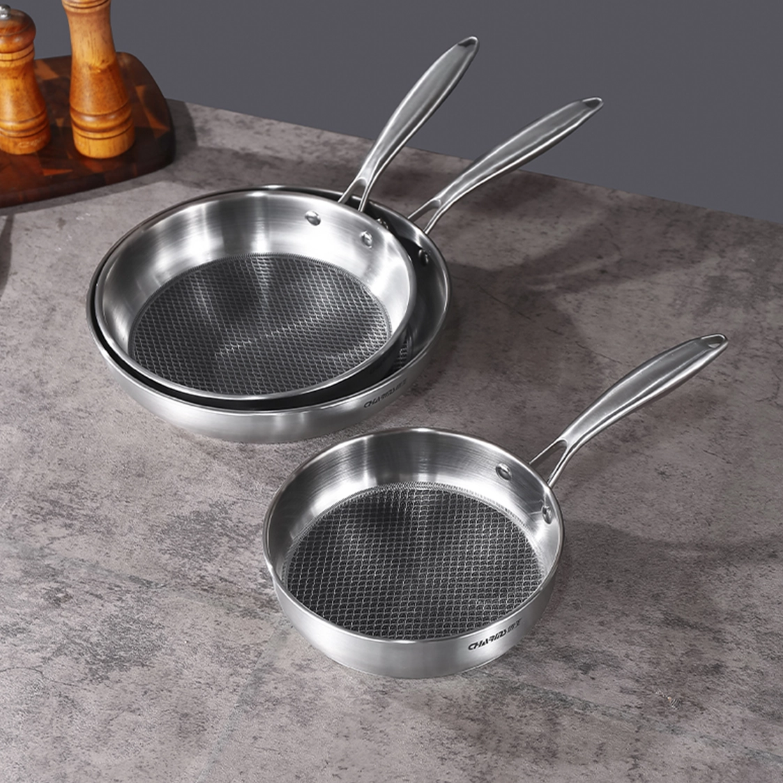 Three shiny stainless steel frying pans positioned on a kitchen counter.