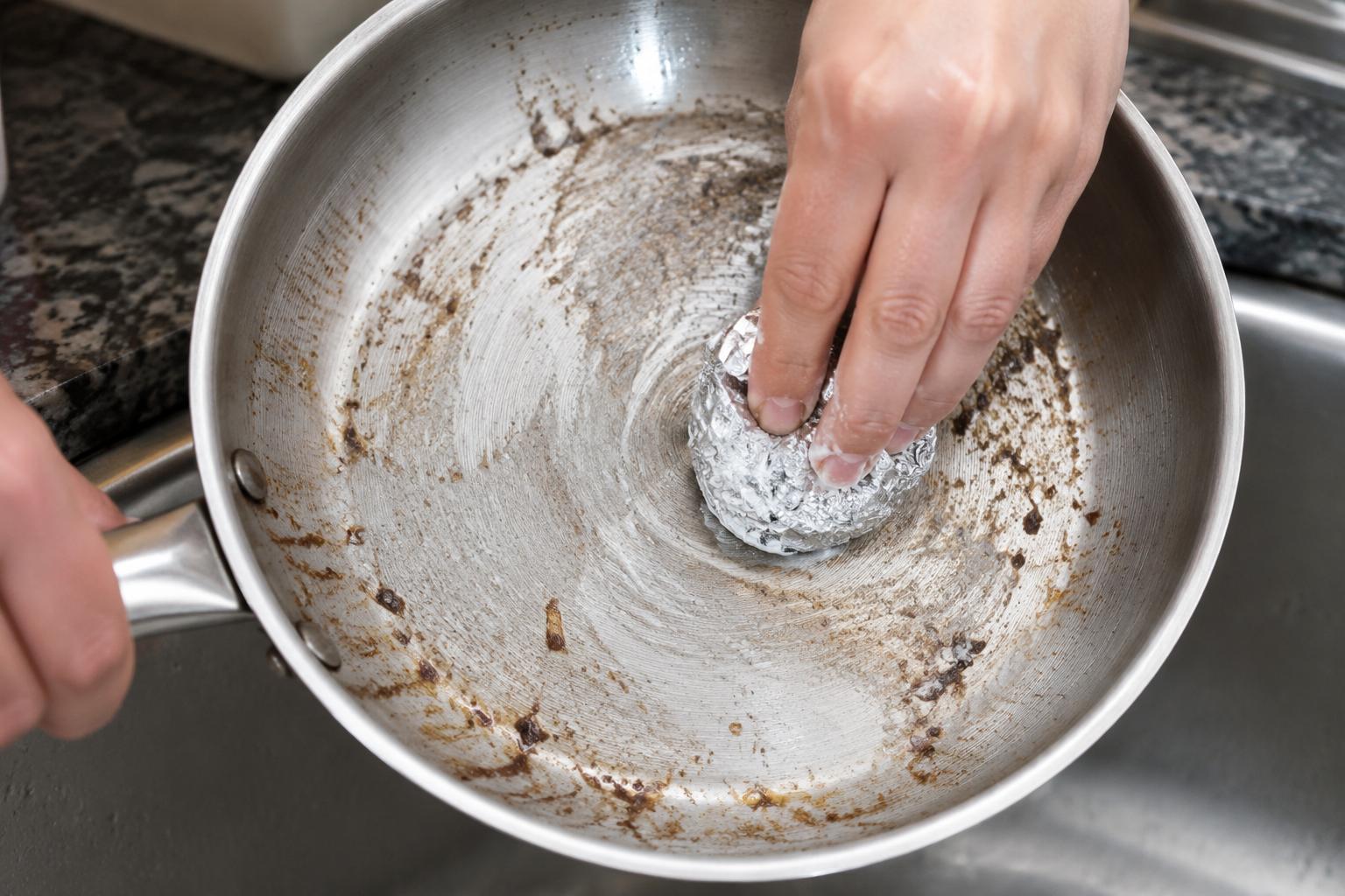 Crumpled aluminum foil ball scrubbing baking soda paste on burnt stainless steel pan - hands-on cleaning action