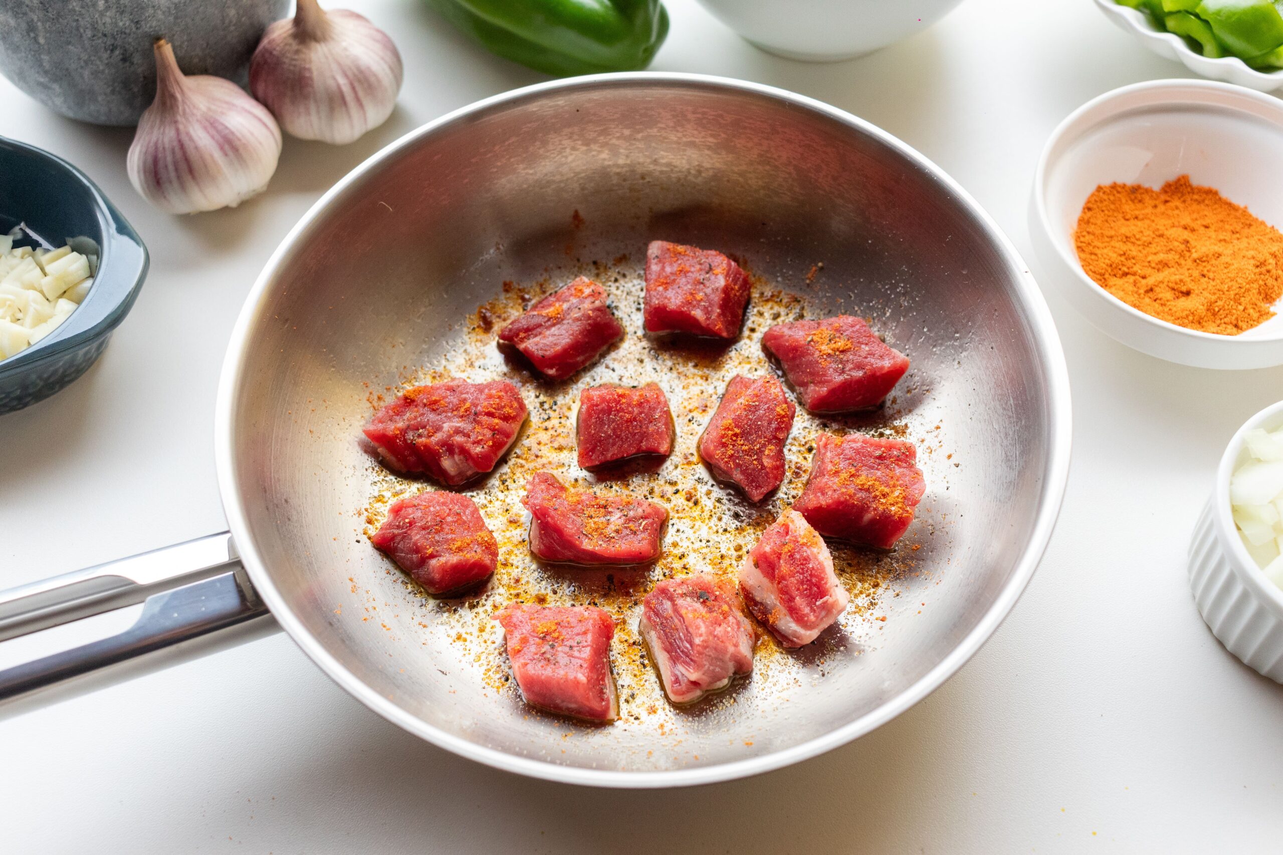Seared seasoned meat in a stainless steel skillet surrounded by fresh herbs and ingredients - showing proper fond development and browning